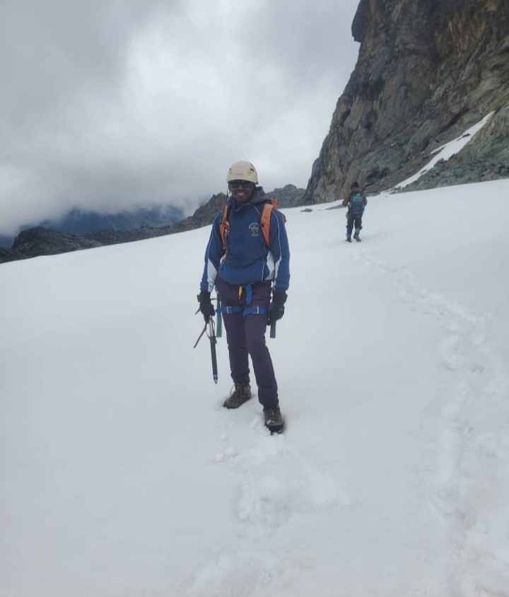 A male hiker standing in deep white snow against a backdrop of rocky glacial peaks on Mount Rwenzori.