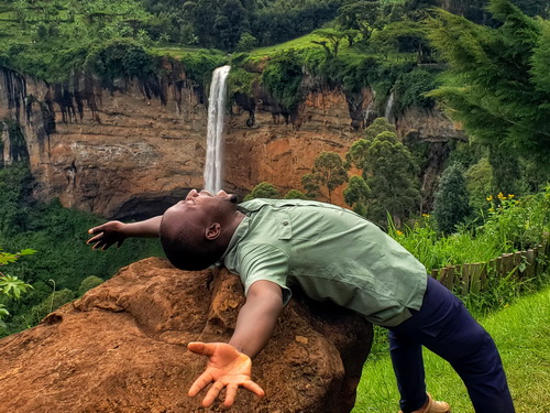 A vertical portrait of a man in a green shirt lying on his back on a reddish-brown rock, arms spread wide and face turned upward toward the sky. In the background, a large waterfall cascades down a rocky cliff face surrounded by terraced green agriculture.. 