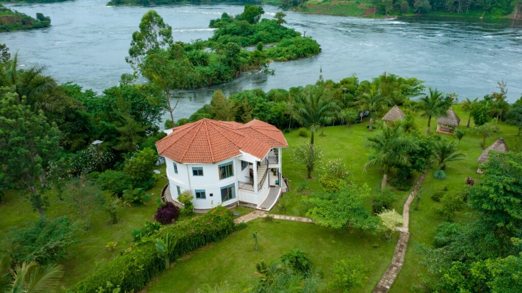Best hotels in Jinja - An aerial view of the white Nile Retreat Luxury Villa with a red tile roof, surrounded by a lush green garden and mature palm trees, directly overlooking the river in Jinja, Uganda.