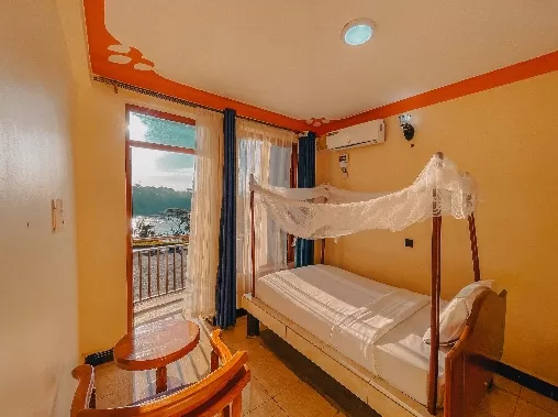 A cozy guest room at Source Garden Hotel in Jinja, featuring a wooden bed with a white mosquito net, warm yellow walls with orange trim, and a private balcony door offering a bright view of the Nile riverfront.