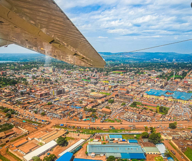 Aerial view of Jinja city from a small plane window showing the urban grid, red dirt roads, and the Nile river in the distance under a blue sky.