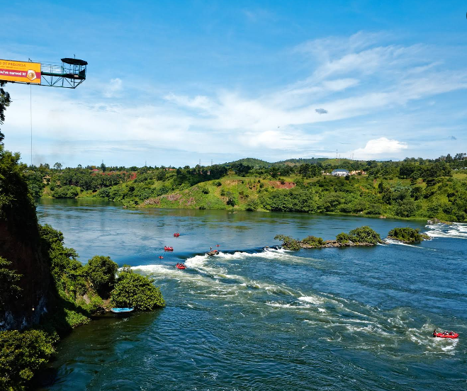 A wide landscape shot of the Nile river in Jinja with a bungee jumping platform on the left and white water rafting boats in the rapids below.