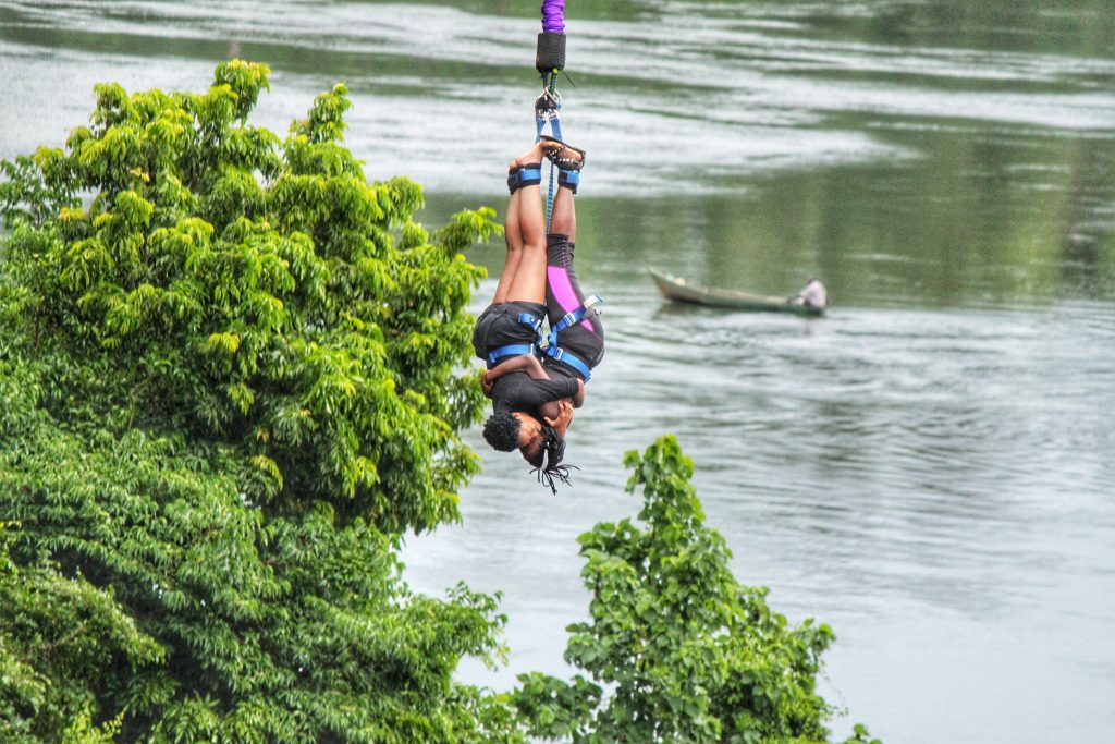 wo people harnessed together performing a tandem bungee jump over the Nile river with green trees and a small boat in the background.