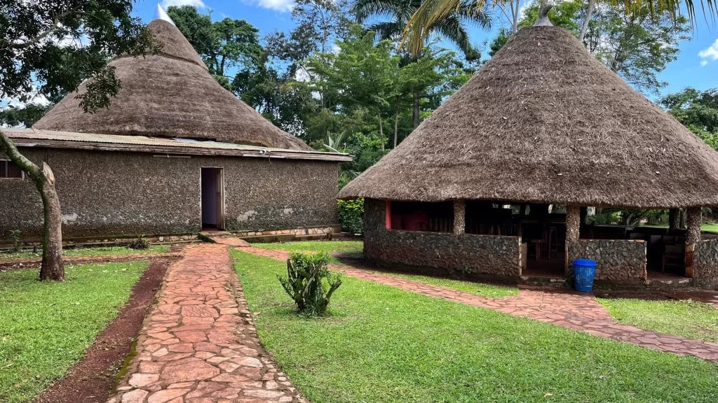 Traditional stone-walled bandas with high, conical thatched roofs at Eden Rock Resorts in Bujjagali, Jinja, connected by red stone-paved walkways and surrounded by green lawns and tropical trees.