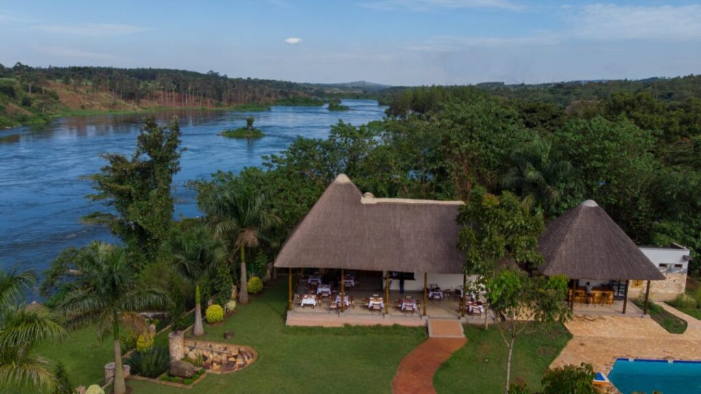 High-angle drone shot of a riverfront resort featuring a large thatched-roof dining hall and a smaller bar pavilion. A red path leads through a green lawn to the main building, which overlooks a wide blue river. A swimming pool is visible in the bottom right corner.