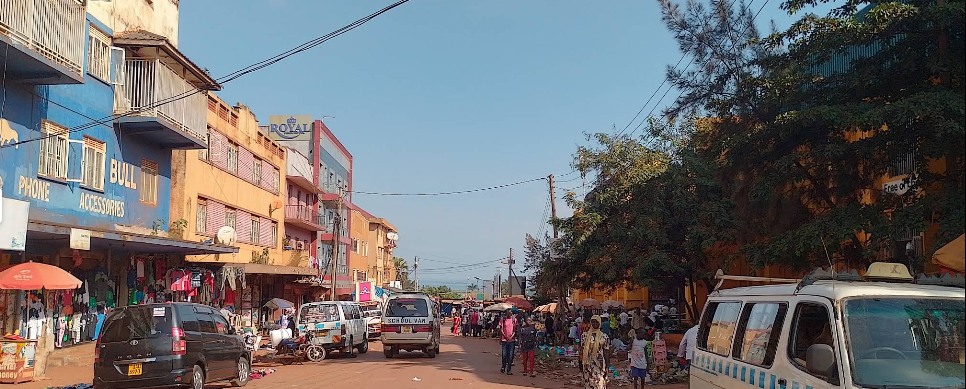 A busy street in Jinja town with parked cars, local shops with blue and yellow facades, and people walking on a sunny day.
