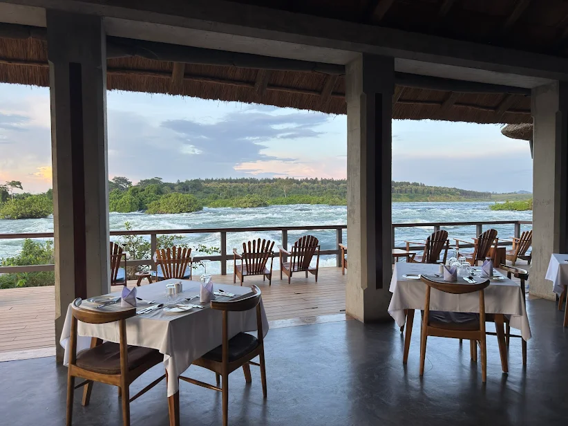 Lemala Wild Waters Lodge Jinja: The Ultimate Private Island Retreat. The main lodge dining pavilion at Lemala Wild Waters, set on a raised timber deck with open-air tables overlooking the Grade 5 rapids of the Victoria Nile during sunset.
