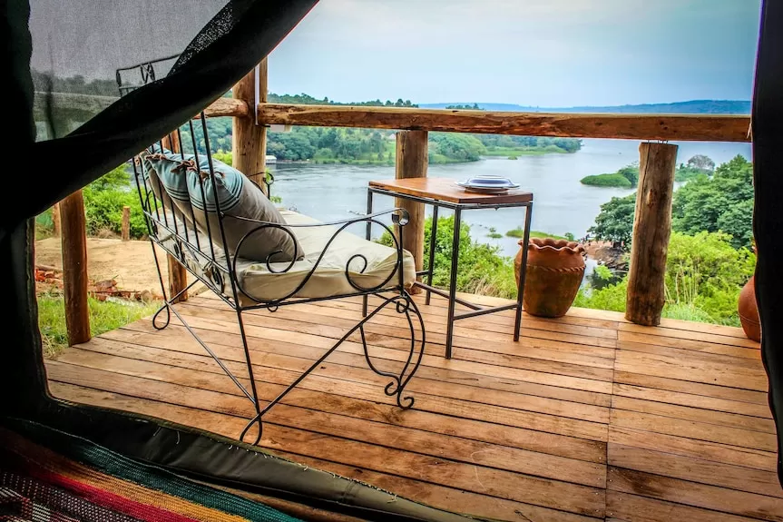 A private wooden balcony at Living Waters Resort in Jinja, featuring a wrought-iron bench with plush cushions, a small wooden table, and a panoramic view of the Nile River and lush green islands.