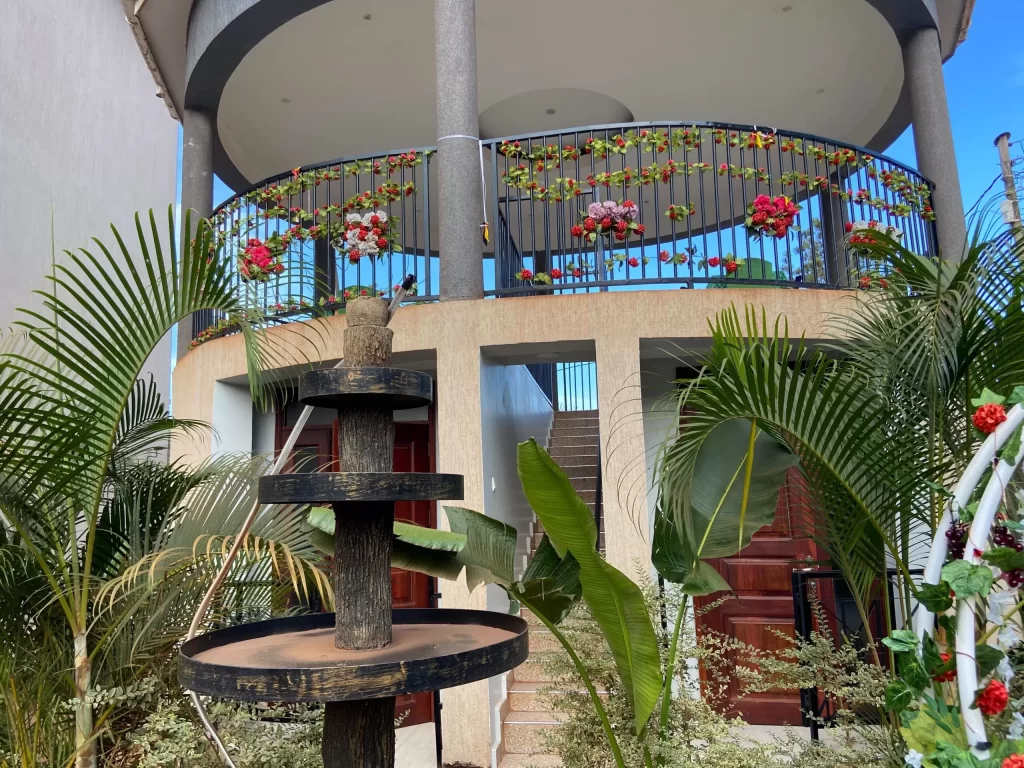 A modern two-story hotel exterior at Spark Africa in Jinja, featuring a curved upper balcony decorated with red and white flowers, a tiered wooden fountain in the foreground, and lush tropical palm plants lining the entrance.