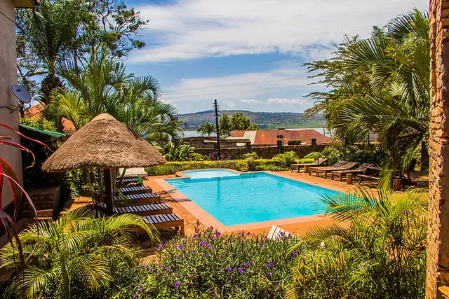 The outdoor swimming pool at Surjios Guest House in Jinja, surrounded by tropical palm trees, thatched umbrellas, and wooden lounge chairs, with a view of the Nile and distant hills in the background.