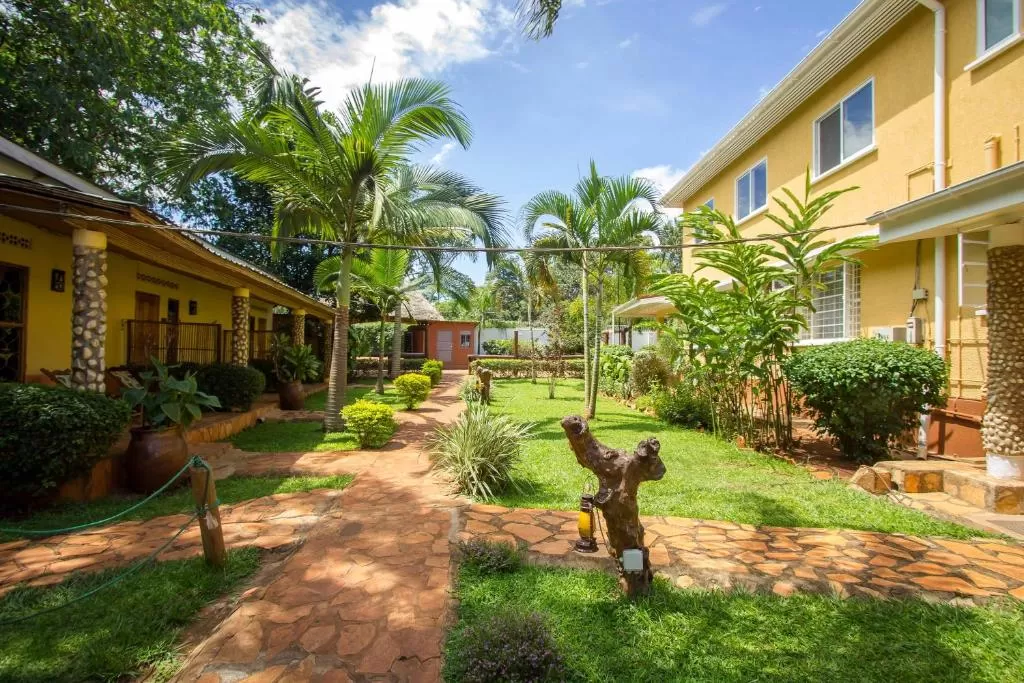 Tropical courtyard at Two Friends Guest House in Jinja, featuring sun-drenched stone walkways, vibrant yellow boutique buildings, and lush green lawns lined with palm trees and potted plants under a clear blue sky.
