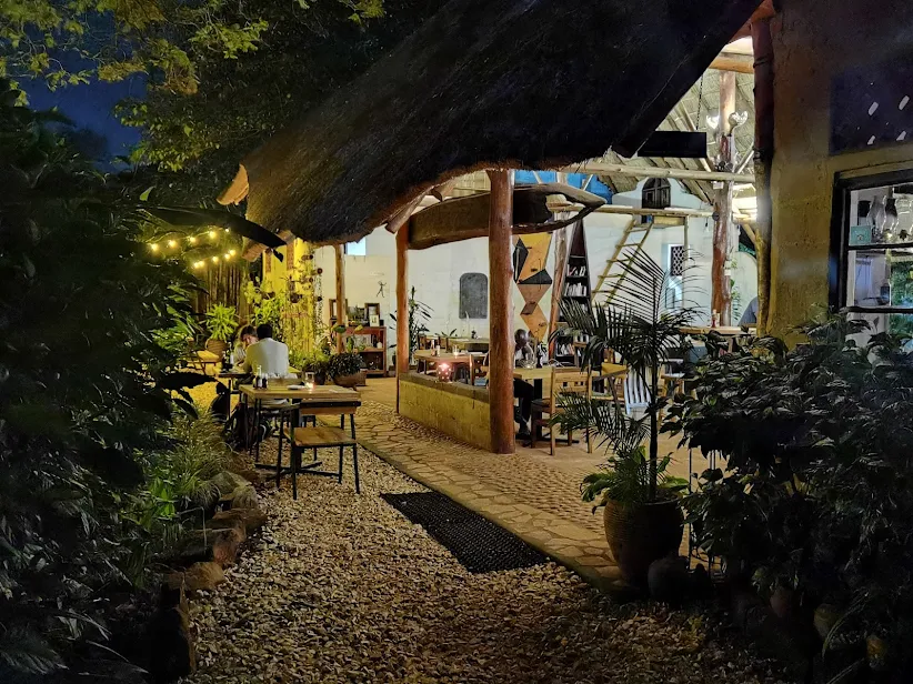 An outdoor evening view of the 30 Wilson garden restaurant, featuring warm string lights, thatched roofing, and guests dining at wooden tables surrounded by lush plants.