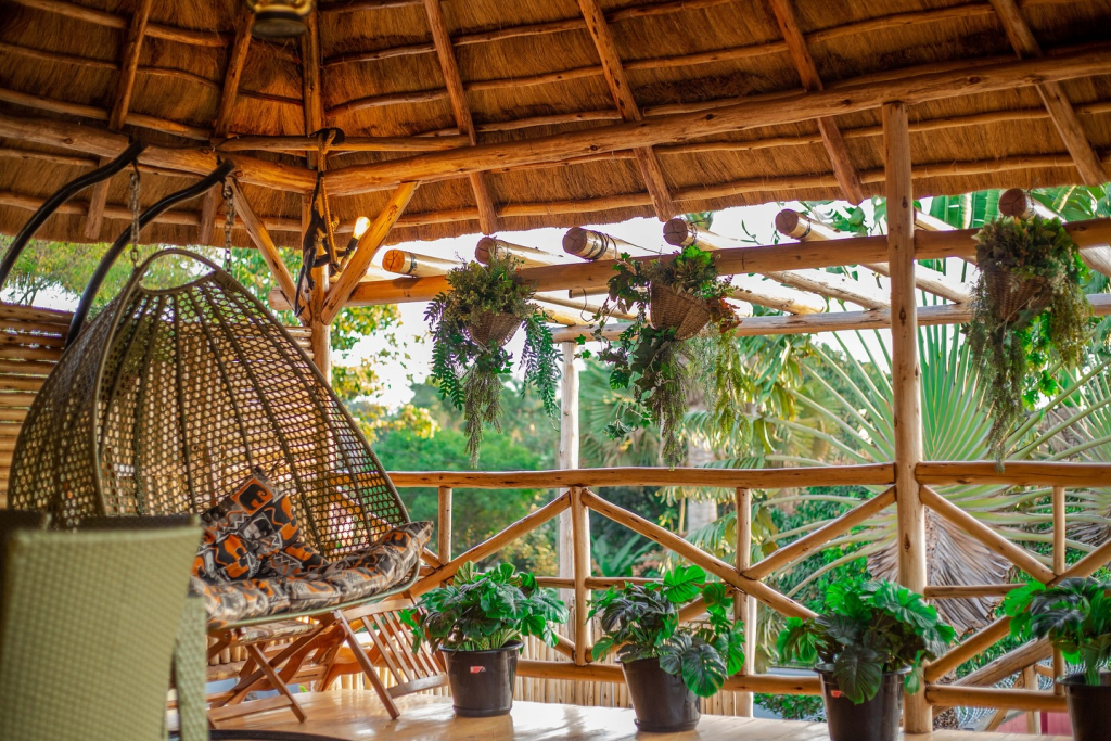 A sunlit wooden balcony at Acacia Nest with a thatched roof, a woven hanging nest chair, and potted greenery overlooking lush tropical trees.