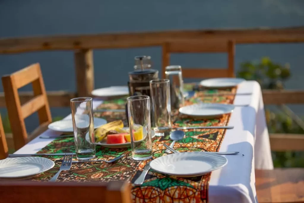 An outdoor breakfast table setting with kitenge fabric runners, fresh fruit plates, and coffee on a wooden deck overlooking the hills in southwestern Uganda.