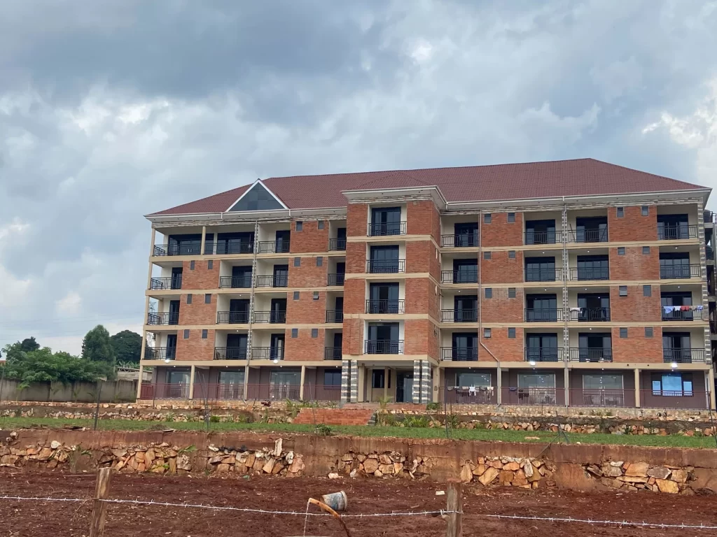 A multi-story red brick hotel complex with dark metal balconies under a cloudy sky, overlooking a large green lawn and tiered stone retaining walls.