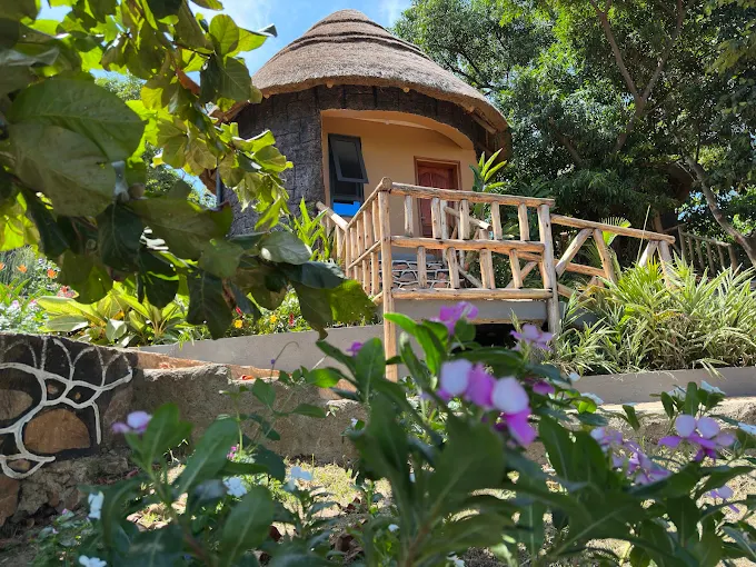 A round, thatched-roof stone cottage at Mountain Inn Resort Kiyindi, surrounded by colorful flowers and lush green trees under a bright blue sky.