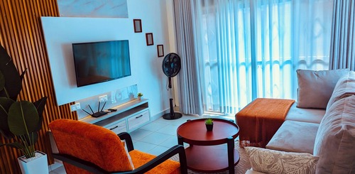 A modern, brightly lit apartment living room featuring an orange armchair, a gray sectional sofa with a rust-colored throw, a flat-screen TV mounted on a white accent wall, and large sheer curtains.