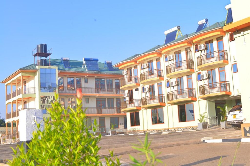 A modern multi-story hotel complex under a clear blue sky with white and yellow walls, orange balcony trim, and solar water heaters mounted on the green rooftops in the where to stay at Nyege Nyege
