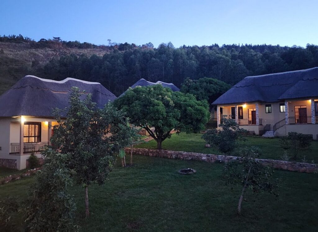An evening view of Stevalyn's Ridge View Lodge featuring thatched-roof cottages with warm porch lights glowing against a dark green hillside and forested ridge under a twilight sky.