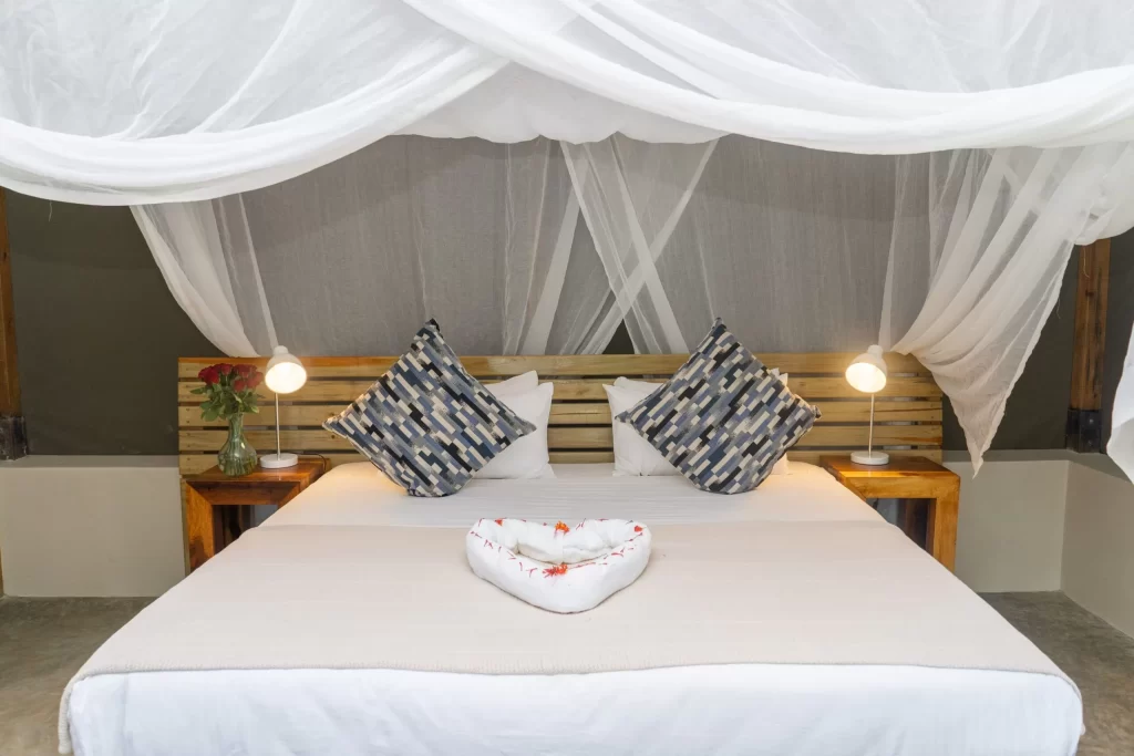 A serene, modern bedroom at Adrift River Club featuring a large bed with white linens, a decorative towel heart, and a wooden headboard. The bed is draped in a romantic white mosquito net and flanked by warm bedside lamps on wooden nightstands.