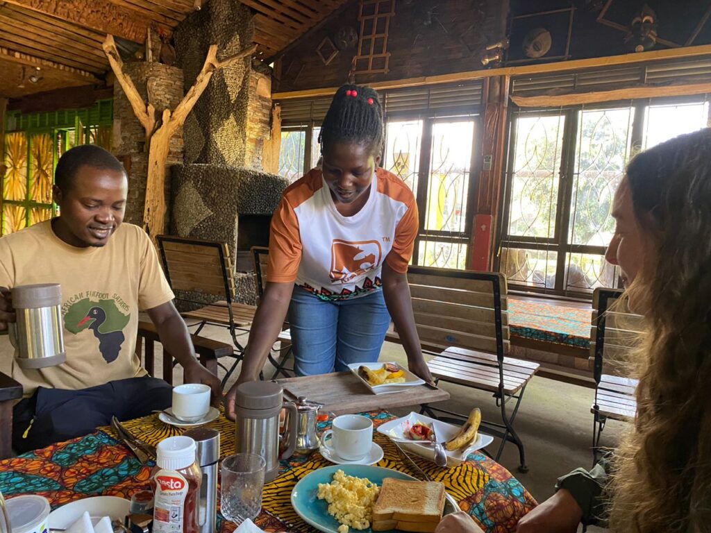A friendly waitress serving plates of fresh breakfast to travelers at a warmly lit wooden table inside a rustic safari lodge restaurant.