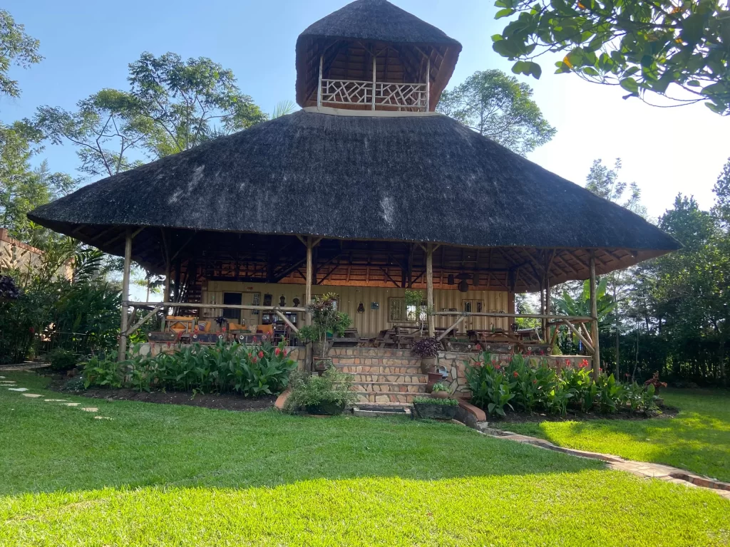 A large, open-air wooden pavilion lounge at Nile It Resort with an expansive two-tiered thatched roof, built on a stone foundation and surrounded by a manicured green lawn and tropical gardens.