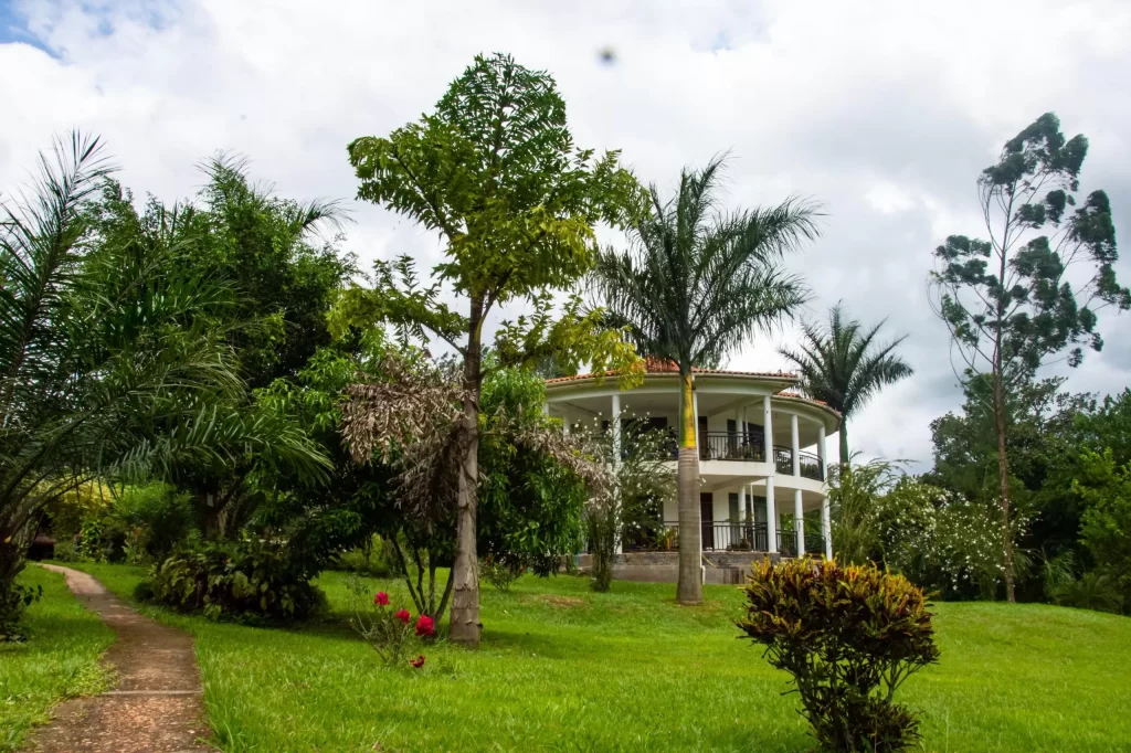 A white, rounded multi-story villa in the "Where to stay at Nyege Nyege guide"with large columns and curved balconies, surrounded by a expansive green lawn, stone pathways, and tall palm trees under a cloudy sky.