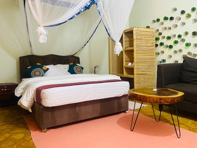 A deluxe guest room featuring a canopy bed with a mosquito net, a slatted timber wardrobe, a solid wood slab coffee table, and the signature creative bottle-art wall at Afro Smiles Hotel & Guest House.