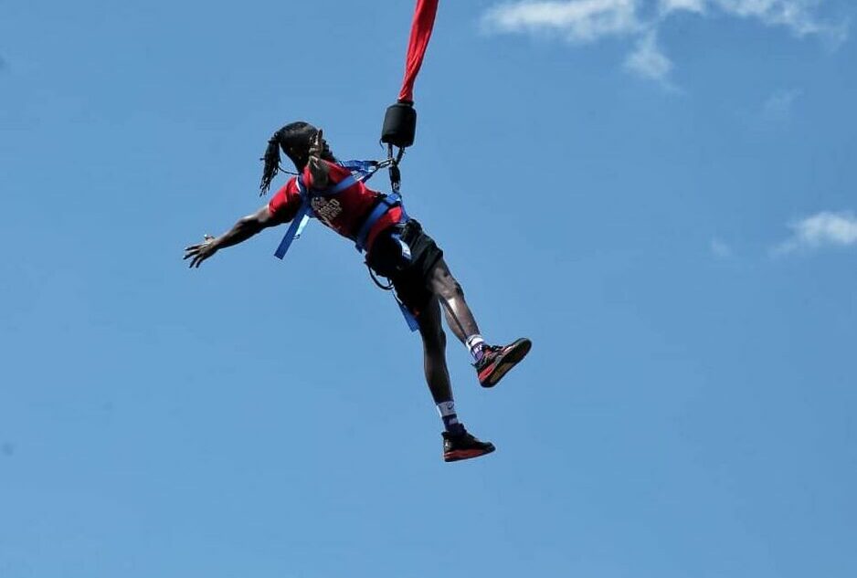 Bungee Jumping in Jinja: The Ultimate Guide to Uganda’s Adventure Capital A thrill-seeker mid-air during a bungee jumping in Jinja experience, wearing a red shirt and secured by a blue body harness attached to a red bungee cord against a bright blue sky