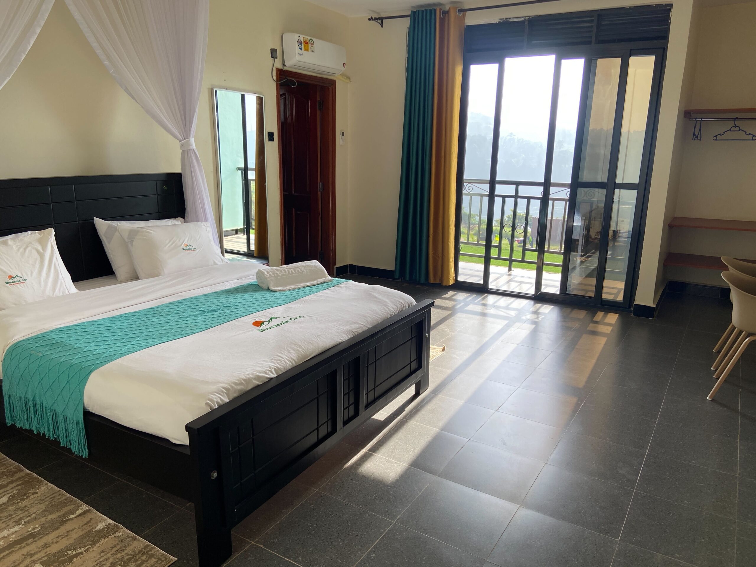 Deluxe room at Mountain Inn Resort Jinja featuring a canopy king bed with teal throws white linen and sliding glass doors opening to a private balcony