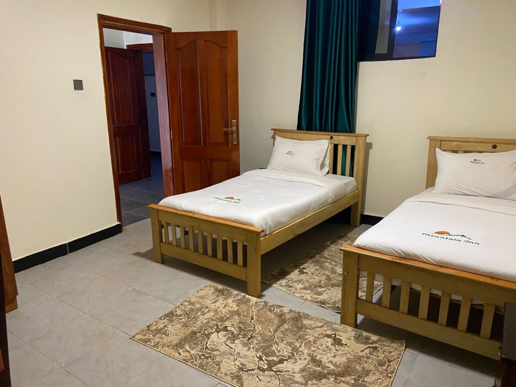 Twin room at Mountain Inn Resort Jinja featuring two separate solid wood single beds with branded white linen tiled floors and a dark teal window curtain