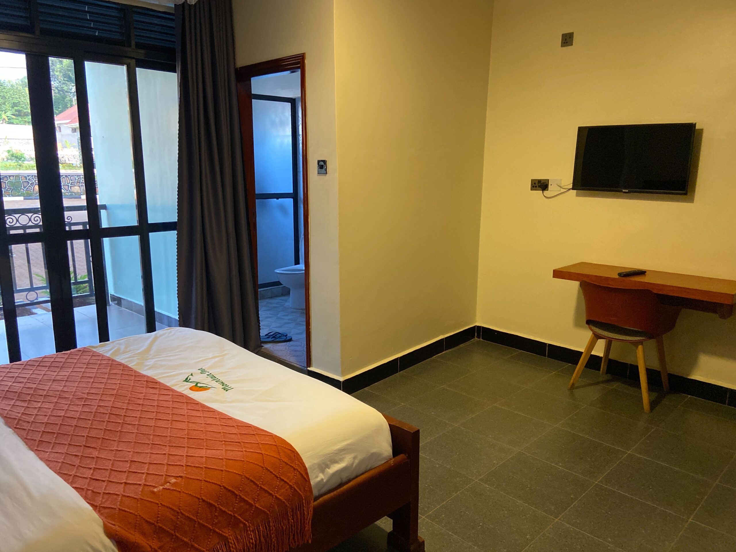 Standard room at Mountain Inn Resort Jinja showing a double bed with an orange throw wall-mounted flat screen TV wooden desk and en-suite bathroom