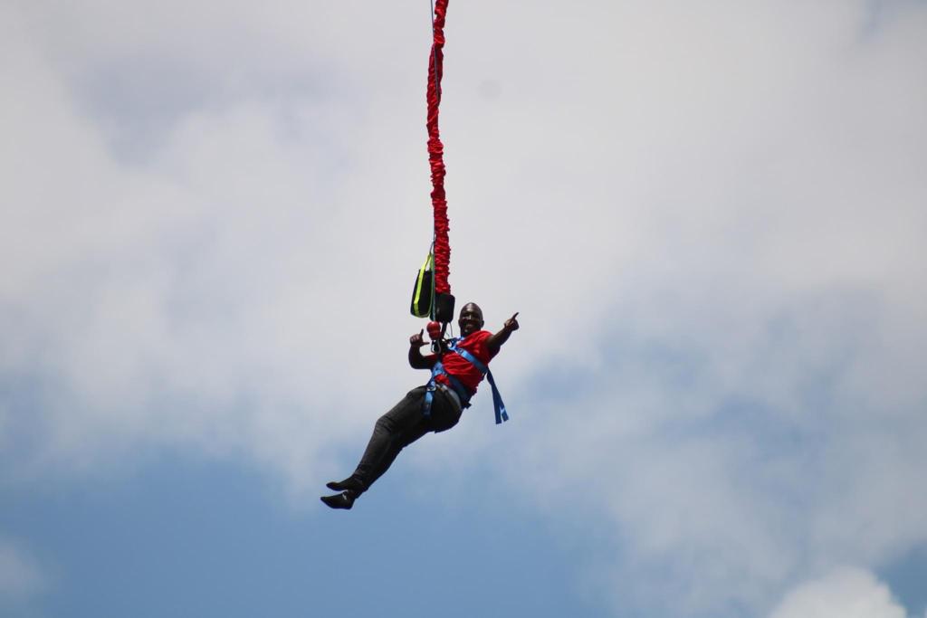 Bungee Jumping in Jinja: The Ultimate Guide to Uganda’s Adventure Capital A smiling jumper gesturing with his hands while suspended in mid-air during a bungee jumping in Jinja experience, wearing a red shirt and secured by a body harness attached to a red bungee cord sleeve against a blue sky with light clouds