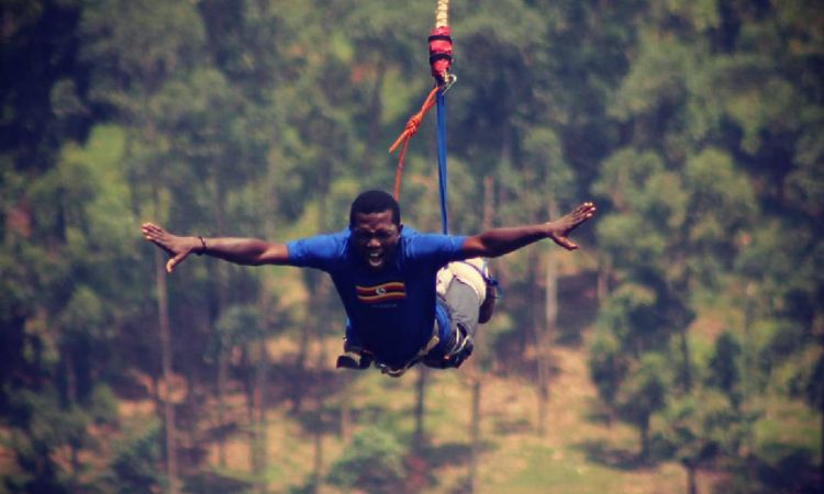 Bungee Jumping in Jinja: The Ultimate Guide to Uganda’s Adventure Capital A man screaming with excitement mid-air during a bungee jumping in Jinja session, wearing a blue shirt featuring the Uganda flag and secured by a full body harness with dense green trees in the background.