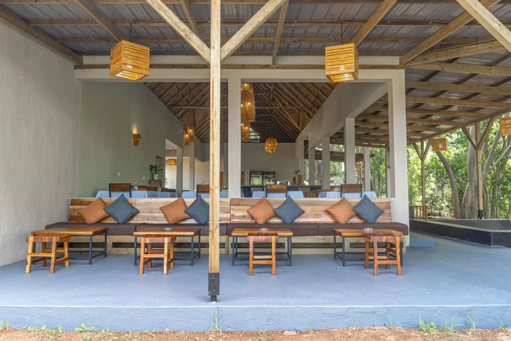 Bungee Jumping in Jinja: The Ultimate Guide to Uganda’s Adventure Capital A covered outdoor terrace lounge at a riverside resort highlighting premium choices for where to stay in jinja, featuring wooden pallet bench seating with blue and orange cushions, low tables, and hanging lantern lights under a rustic wooden frame structure