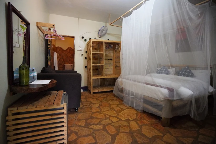 A cozy guest room interior showing local stone flooring, a timber canopy bed with a draped mosquito net, a slatted wooden wardrobe, and honest handcrafted details at Afro Smiles Hotel & Guest House.