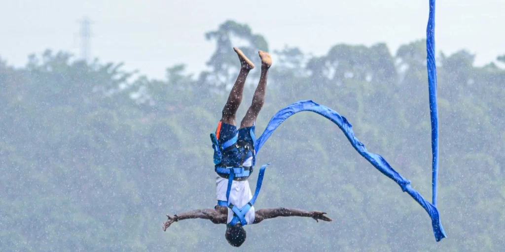 Bungee Jumping in Jinja: The Ultimate Guide to Uganda’s Adventure Capital A jumper falling upside down with arms outstretched wide during a bungee jumping in Jinja session in light rain, secured by a blue body harness attached to a trailing blue bungee cord against a background of misty green trees.