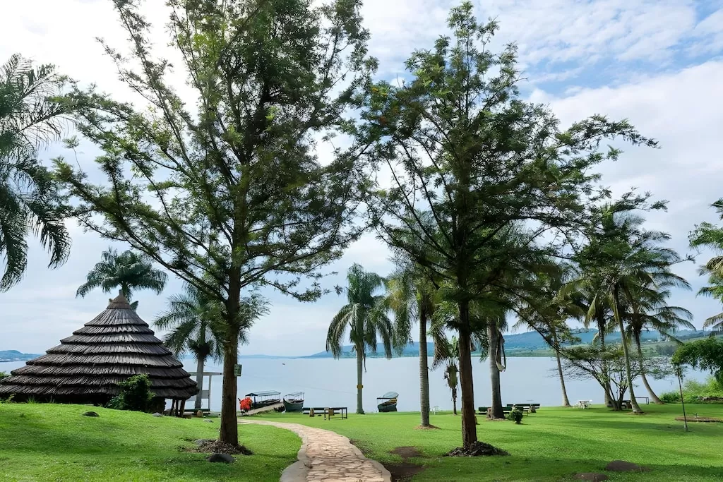 A stone brick pathway curves through a green grassy lawn with palm trees and a traditional thatched roof hut leading toward the River Nile in Jinja Uganda.
