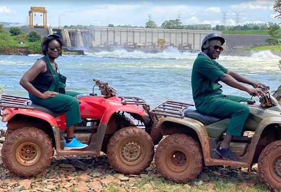Two travelers wearing green protective overalls sit on their ATVs along the rocky banks of the River Nile with a hydroelectric power dam structure in the background, showing the landmarks visited during quad biking in jinja uganda.