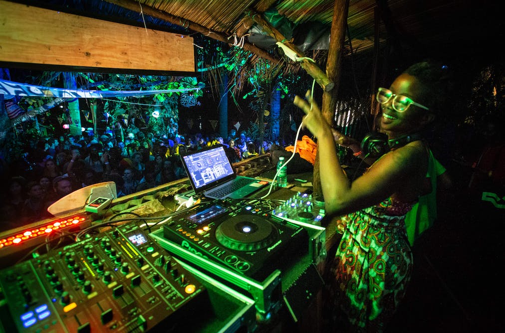 A female DJ with glasses and a patterned dress performing at a thatched DJ booth surrounded by jungle greenery and neon lights in front of a night crowd at Nyege Nyege festival in Jinja Uganda.