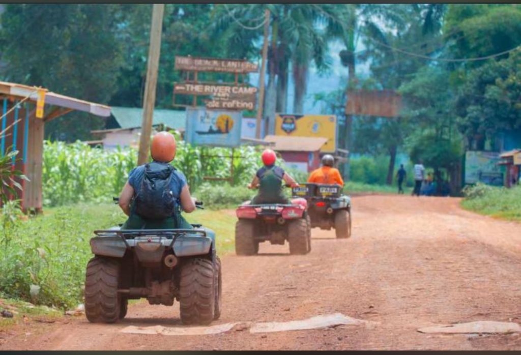 Three travelers riding ATVs down a red dirt road past local adventure signposts, showcasing the rural village routes included in quad biking in Jinja Uganda.