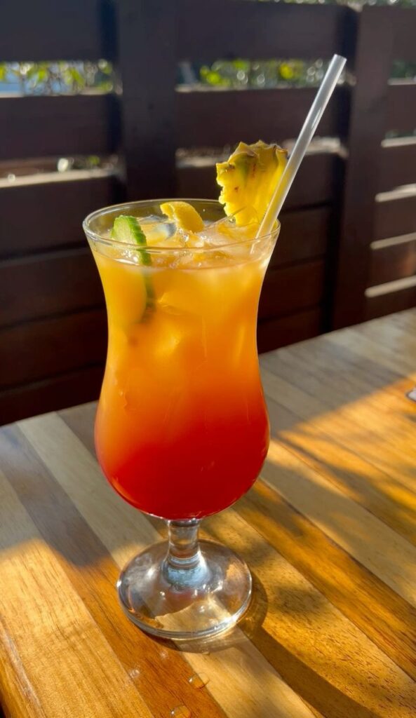 A layered red and yellow tropical sunrise cocktail in a hurricane glass garnished with a pineapple wedge, lime slice, and straw on a wooden table in Jinja Uganda.