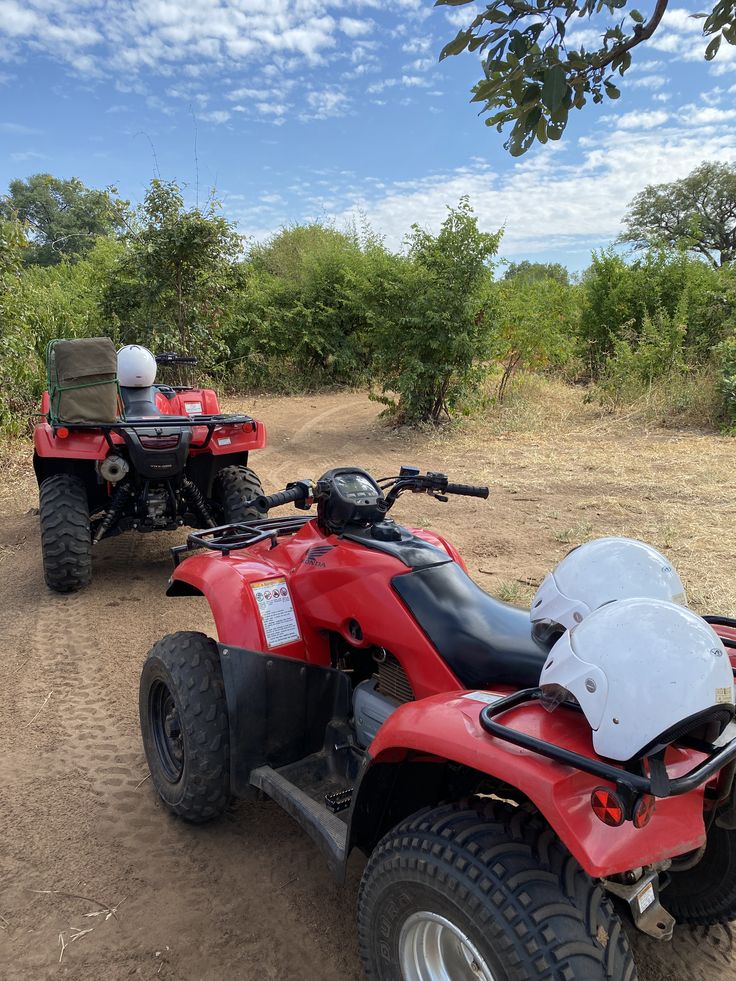 Two red Honda quad bikes parked on a dirt track in a grassy, bush terrain under a blue sky, with white safety helmets resting on the rear rack, highlighting equipment logistics for quad biking in Jinja Uganda.