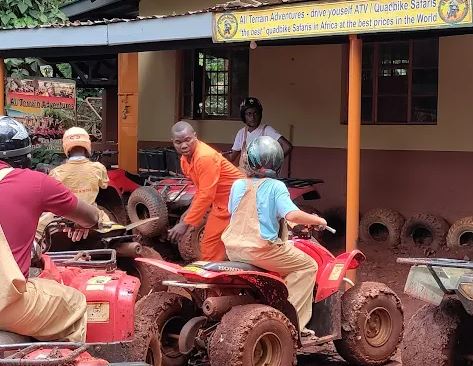 A group of riders in helmets and protective overalls receiving a safety briefing next to mud-covered ATVs at an adventure base camp for quad biking in Jinja Uganda.