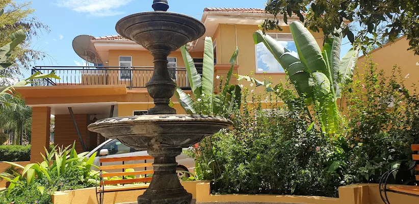 A two-tier stone water fountain in the foreground of a multi-story yellow luxury resort villa surrounded by lush green tropical plants and banana leaves, illustrating where to stay in Jinja Uganda.