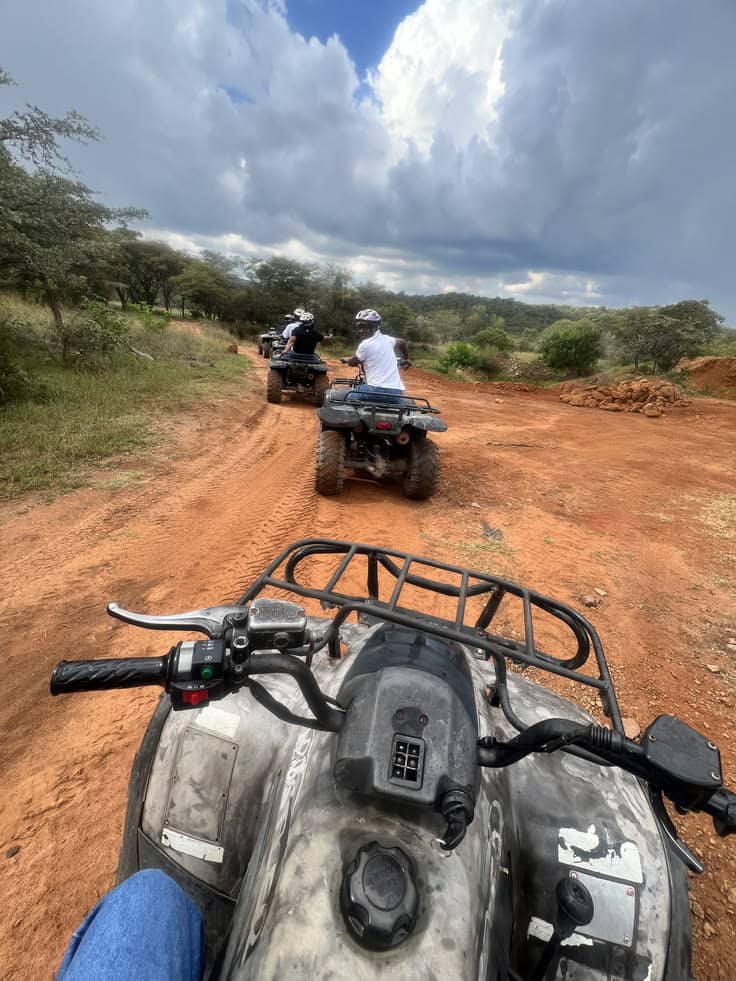 A first-person POV shot of riding an ATV quad bike on a dirt trail, following a group of riders through red clay paths and greenery under a cloudy sky in Jinja Uganda.
